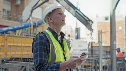 Man builder in hardhat and highvis vest holds smartphone and looks up at bu.. Stock Footage 339178050