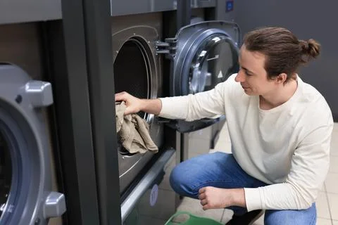 Man with a bun crouching to load clothes into a washing machine at a self-s.. Stock-Fotos