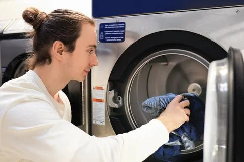 Man with bun loading clothes into washing machine at public laundromat. Stock Photos