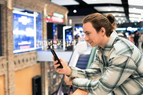 Man with bun using smartphone and smiling in shopping mall with blurred lig.. Stock Photos