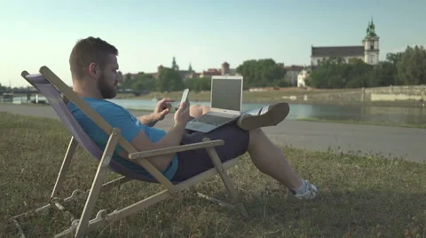 Man calls by smartphone, browsing laptop, lying on sunbed by the river. Vídeo Stock 65205356