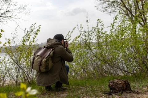 Man with a camera sits in thickets of grass near the river Stock Photos