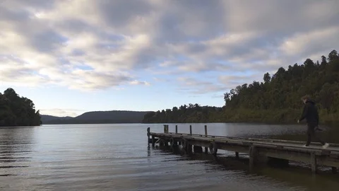 Man with camera walking towards the end of tranquil Mapourika lakeside jetty Stock Footage 119152128
