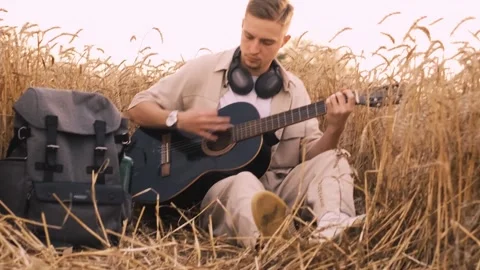 Man camper sitting on grass in field at sunset playing guitar singing song looki Stock Footage 250623682