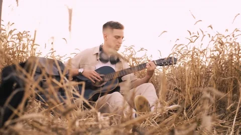 Man camper sitting on grass in field at sunset playing guitar singing song looki Stock Footage 250623692