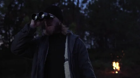 Man camping in a forest looking with binoculars in front of a campfire. Stock Footage 76473463