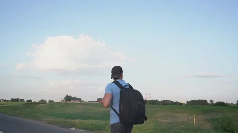 Man with cap and backpack walks along center of road near village Stock Footage 166172361