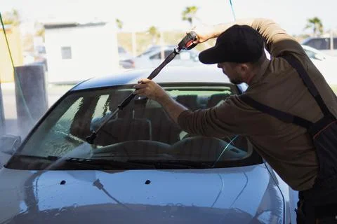 Man in cap and work overalls carefully washing a car windshield with a high.. Stock Photos
