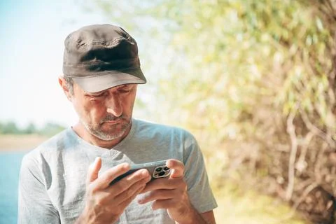 Man in a cap looking down thoughtfully at a smartphone while standing outdo.. Stock Photos