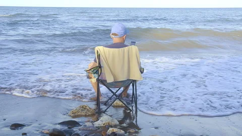 Man in a cap sits on the beach on a beach chair reading a message in his phone Stock Footage 274164115
