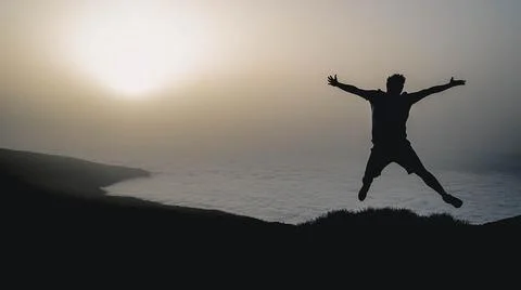 A man is captured in mid-jump on a cliff overlooking a serene body of water a Fotos de archivo