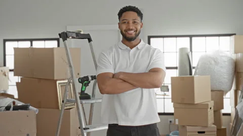 Man with cardboard boxes and cordless drill, arms crossed beside ladder in .. Stock Footage 337438751