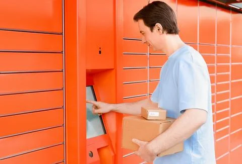 Man with cardboard boxes uses the touch screen of an automated orange parce.. Stock Photos