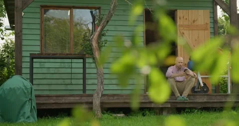 A man carpenter is sitting on the veranda of his tiny carpentry workshop resting Stock Footage 251798210