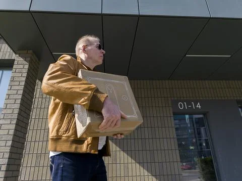 Man carries cardboard parcel while walking past modern building facade. Perso Stock Photos