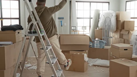 Man carrying boxes and stepping past a ladder, raising arm while navigating.. Stock Footage 337390728
