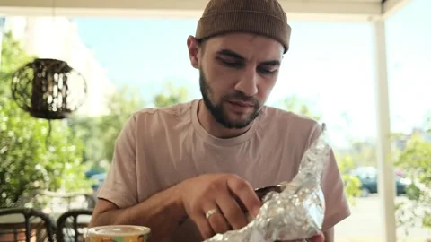A man in casual clothes opens a foil-wrapped burrito while sitting in an outdoor Stock Footage 342544994
