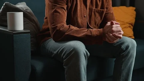 A man in casual clothing sits on a couch with his hands clasped together. He Stock-Footage 339097553