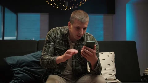 Man celebration victory using phone sitting at couch indoor interior.Young guy Stock Footage 225656235