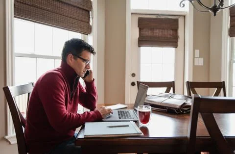 Man on cellphone working from home using a computer at a dining table. Foto stock