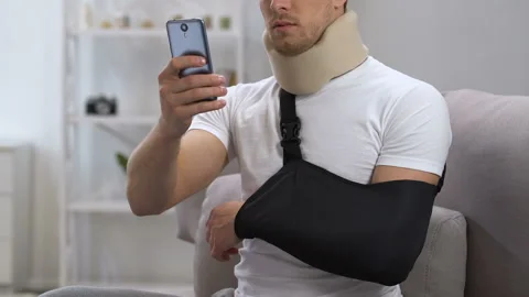 Man in cervical collar and arm sling chatting smartphone at home, rehabilitation Stock Footage 107747459