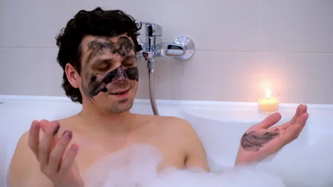 Man with charcoal face mask lying in bath with foam candle meditating at home. Stock Footage 270540277