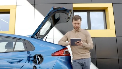 Man is charging an electric car while standing at a station,holding tablet Stock Footage 291879135