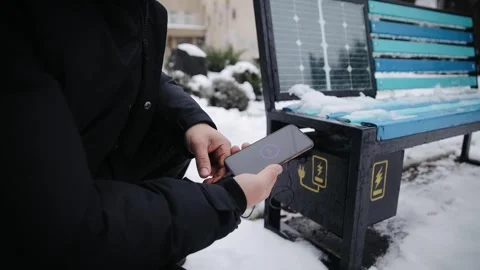 Man Charging Mobile Phone via USB Outdoors in winter. Public Charging on Bench Stock Footage 258679641