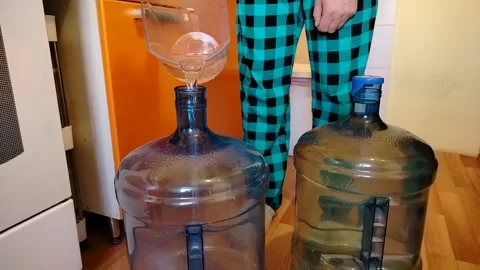 A man in checkered pants in the kitchen pouring water into 2 large bottles Stock Footage 308574816