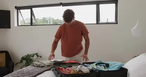 Man checking list and folding garments into suitcase in bedroom packing for trip Stock Footage 316478674