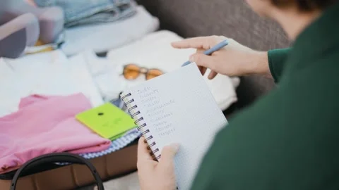 Man checking a packing list in a notebook while packing a suitcase for summer va Stock Footage 339273571