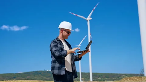 Man checks data on laptop at wind farm during sunny day. A man in a safety Stock Footage 342736901