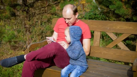 A man with a child using a smartphone sitting on a bench. Father and son smiling Stock Footage 97637123