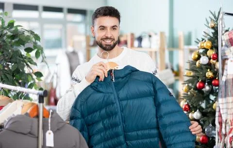Man chooses a down jacket against the background of a Christmas tree in a store Stock Photos