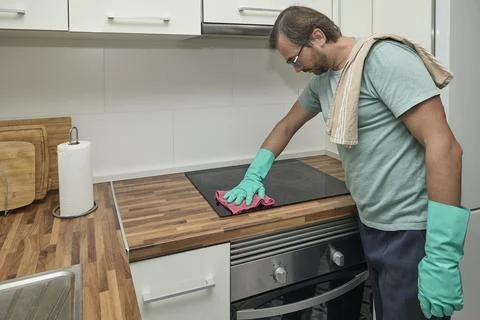 Man cleaning electric stove in modern kitchen with gloves and sponge Stock Photos