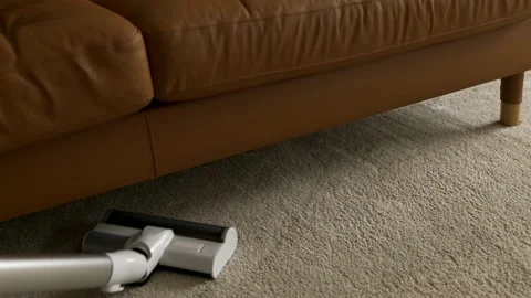 A man cleans the surface of a carpet under the sofa with a cordless vacuum 스톡 동영상 202010132