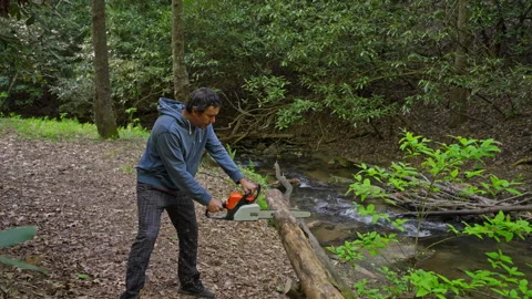 Man clearing a fallen log over a stream with a chainsaw in a wooded area Video stock 320920476