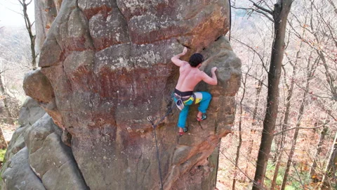 Man climbs high rock wall surrounded by bare autumn trees, seen from drone Stock Footage 346768203