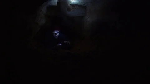 Man climbs outward from hole inside cave with flashlight and smartphone in hands Stock Footage 80062868