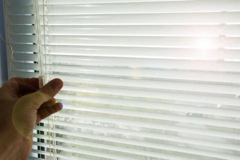 Man closes or opens lowered horizontal white aluminum blinds. Through the ope 스톡 사진