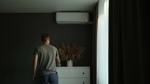 Man closes window, takes remote control, turns on air conditioner. 動画素材 212746503