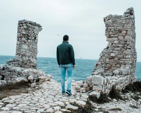 Man on Cobblestone Path Between Stone Ruins Facing the Ocean Stock Photos