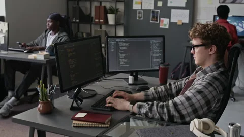Man Coding on Dual Monitors with Multiethnic Team in Tech Office Stock Footage 336712694