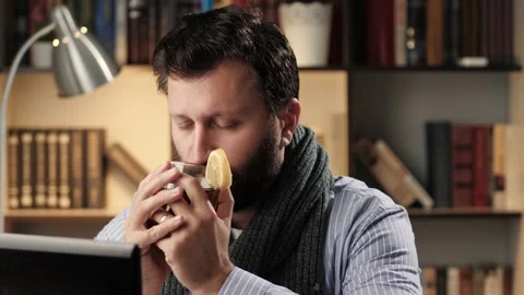 Man with cold is slowly drinking hot tea with lemon at workplace in office or Stock Footage 140483515