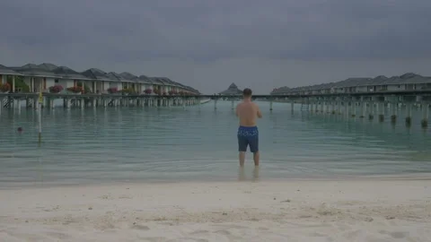 A man comes into the ocean, standing in front of water villas. White sandy beach Stock Footage 90290160