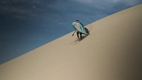 Man coming down a large dune in desert with stand up paddle board in his hands. Stock Footage 244605175