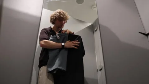Man compares different clothes and chooses an outfit inside a fitting room Stock Footage 344856451