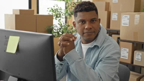 Man at computer clasping hands and smiling among stacked parcels and shippi.. Stockbeeldmateriaal 337369865