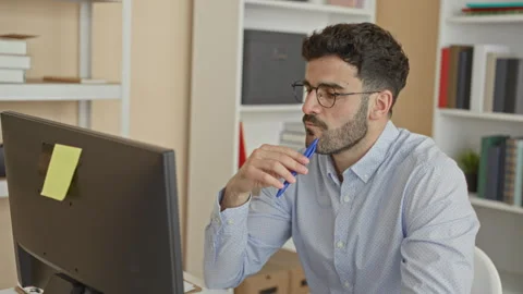 Man at computer holds pen to lips while studying the monitor in an office b.. Stock Footage 337373408