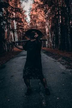 A man in a conical hat and mask stands on a road in the forest, holding a k.. Stock Photos
